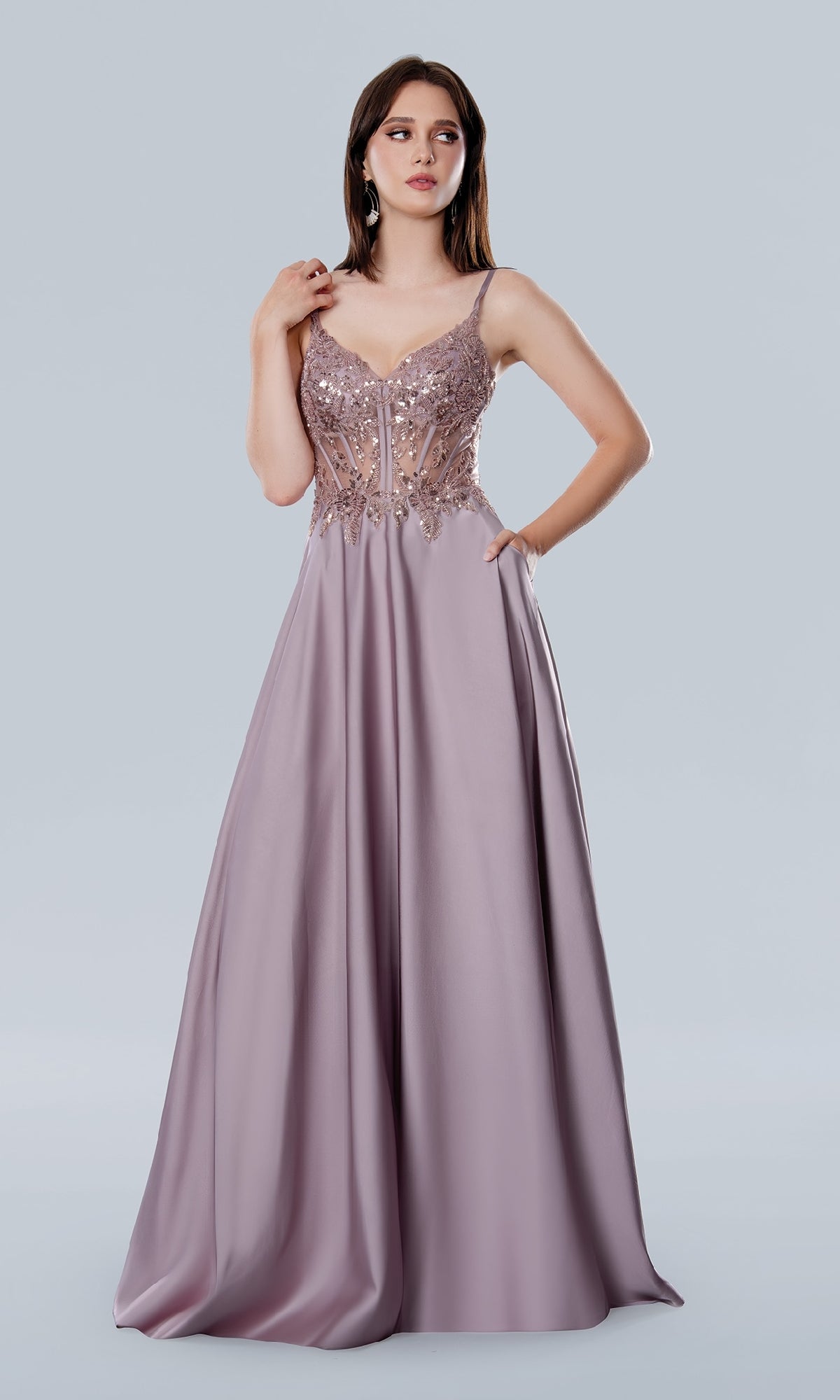A woman models the Long Prom Dress: Stella Couture 25159—a floor-length, sleeveless lavender gown with a beaded semi-sheer bodice and flowing satin skirt, set against a plain light background.